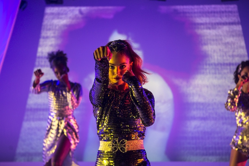 Women in bedazzled silver costumes have their fists in front of their faces like boxers. A bright purple background augments their costumes.