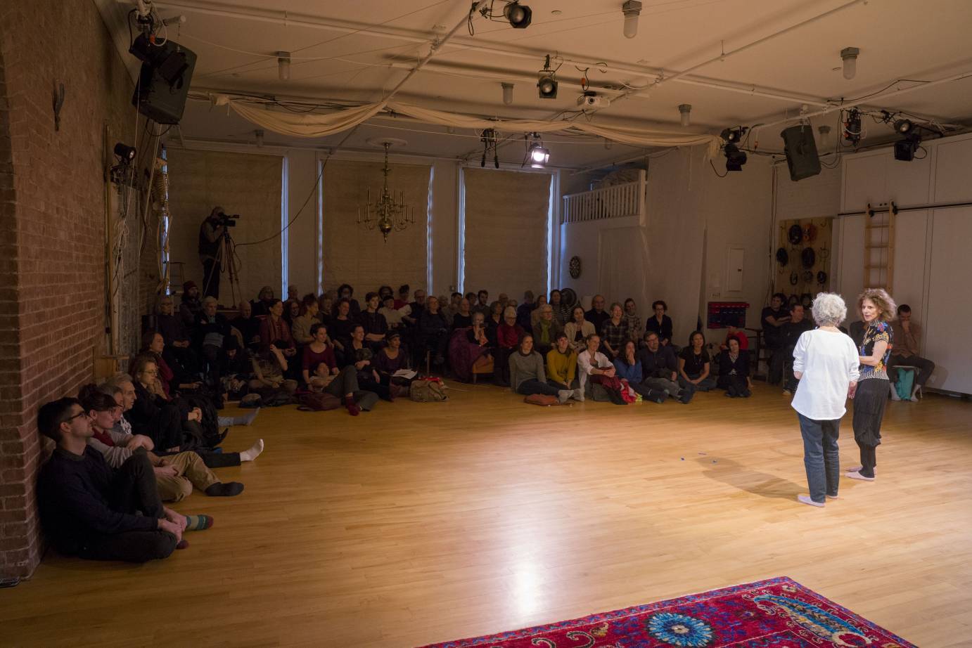 Simone Forti and Cathy Weis stand on the WeisAcre loft stage in front of a seated audience. 