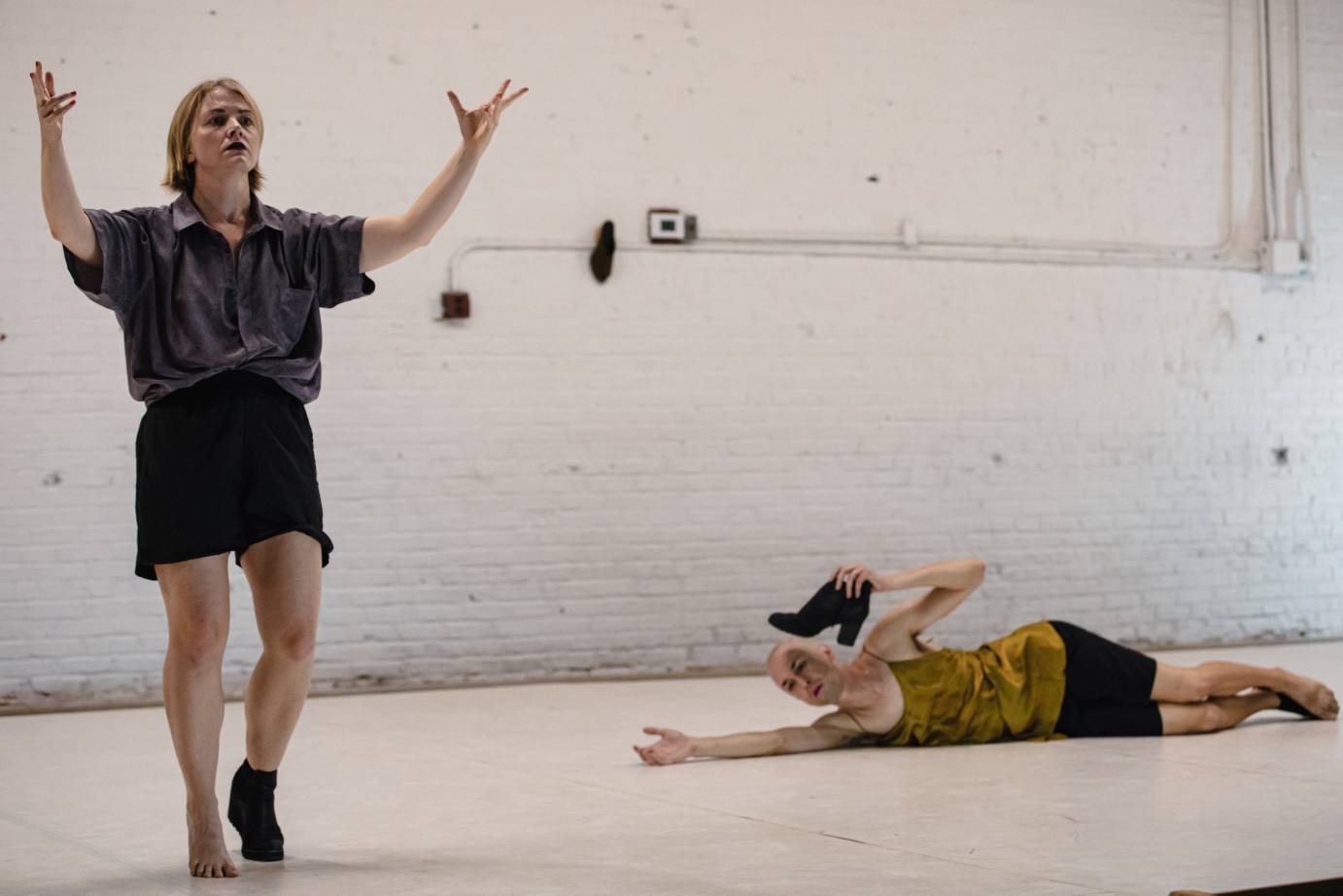 One woman standing facing forward with arms outstretched, splayed fingers facing upward, wearing one black boot (the other foot is barefoot), in contrast with another woman lying on the ground holding the standing woman's boot.