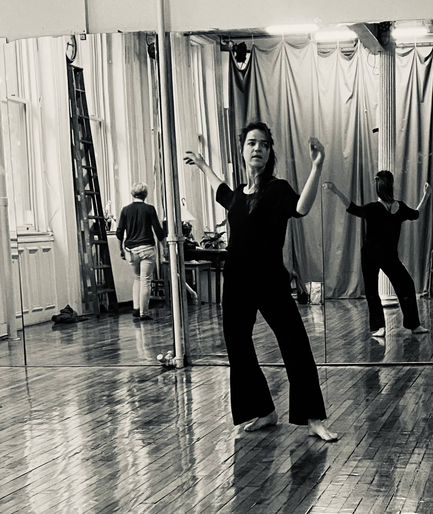 white woman dancer dressed in black facing forward confidently in an open 4th position. The back of a man retreating is to the left of the photo.