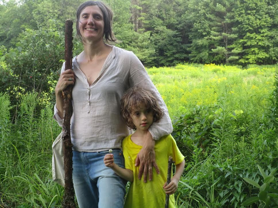 A white woman with a broad smile dressed in a loose long-sleeved shirt and jeans drapes on arm over a young boy with floppy blond hair wearing a lime green t-shirt. They are standing in a green field surrounded by forest.