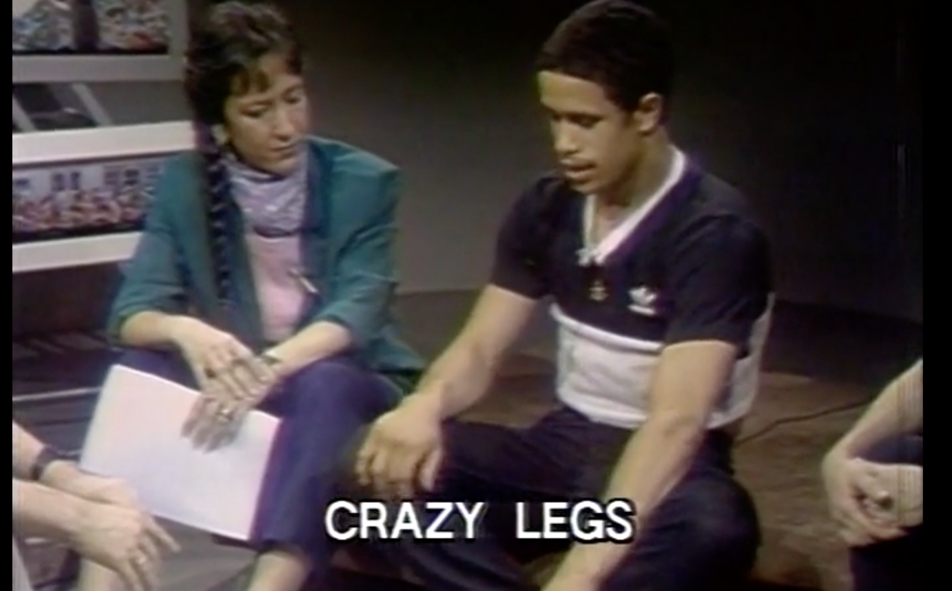 interviewer white colored woman in green jacket sits on floor holding her notes and listening to a light carmel colored man who is a b-boy dancer talking about his art
