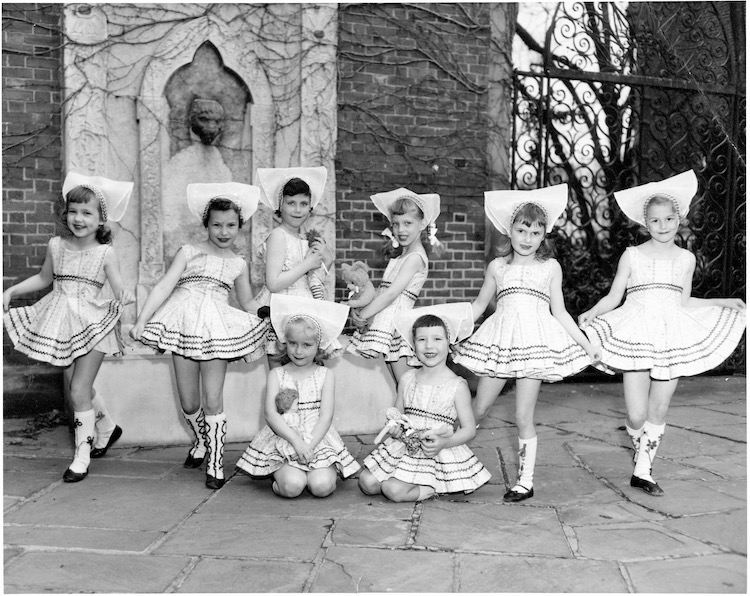 Black and white photo of a group of young fair skinned girl children, in white identically decorated dresses with short full skirts, with a stiff white headress, black ballet shoes and white decorated knee-socks, some of the girls hold dolls