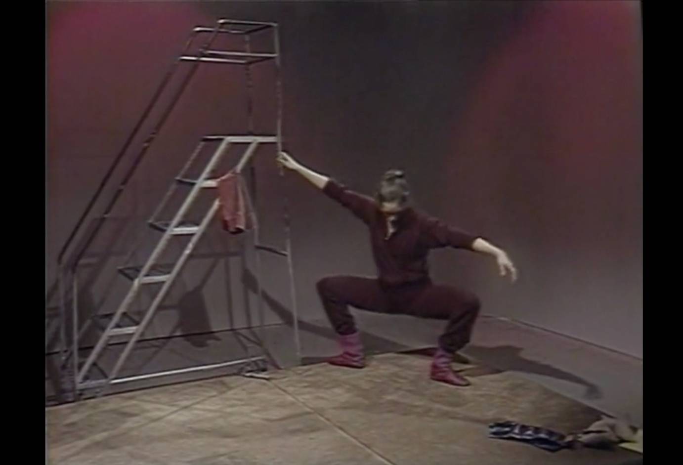 a ballerina, white woman with light hair up in a bun, holds on to a stage ladder, as she performs a deep knee bend. the walls behind her are a light burgundy and her warm up suit is a dark burgundy she is wearing a lighter colored dance warm up boot