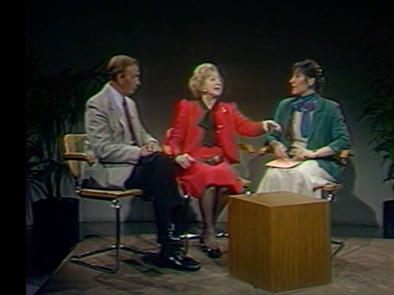 an interview, 2 legendary older ballet dancers are interviewed. Man is white light jacket, dark pants tie, woman is in bright red suit jacket and dark top, the interview a light skinned woman with dark hair is wearing a green jacket over a white dress with blue scarf