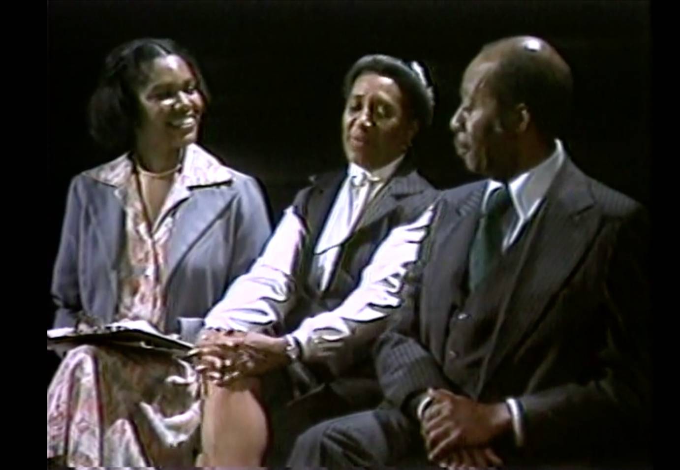 a young  smiling black woman in a white dress with purple flowers and purple jacket interviews two elder black performers of the Lindy Hop, one woman and one man, both elders  dressed formally in black and white,  in a dark =walled studio 