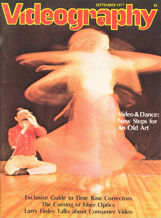 Cover of a 1977 Videography Mag. man with dark hair, orange-red long sleeved buttondown shirt, yellow jeans and black socks, sits on floor filming female dancer, who because she is twirling looks like a whirle of orange and white light  face covered by a camera, 