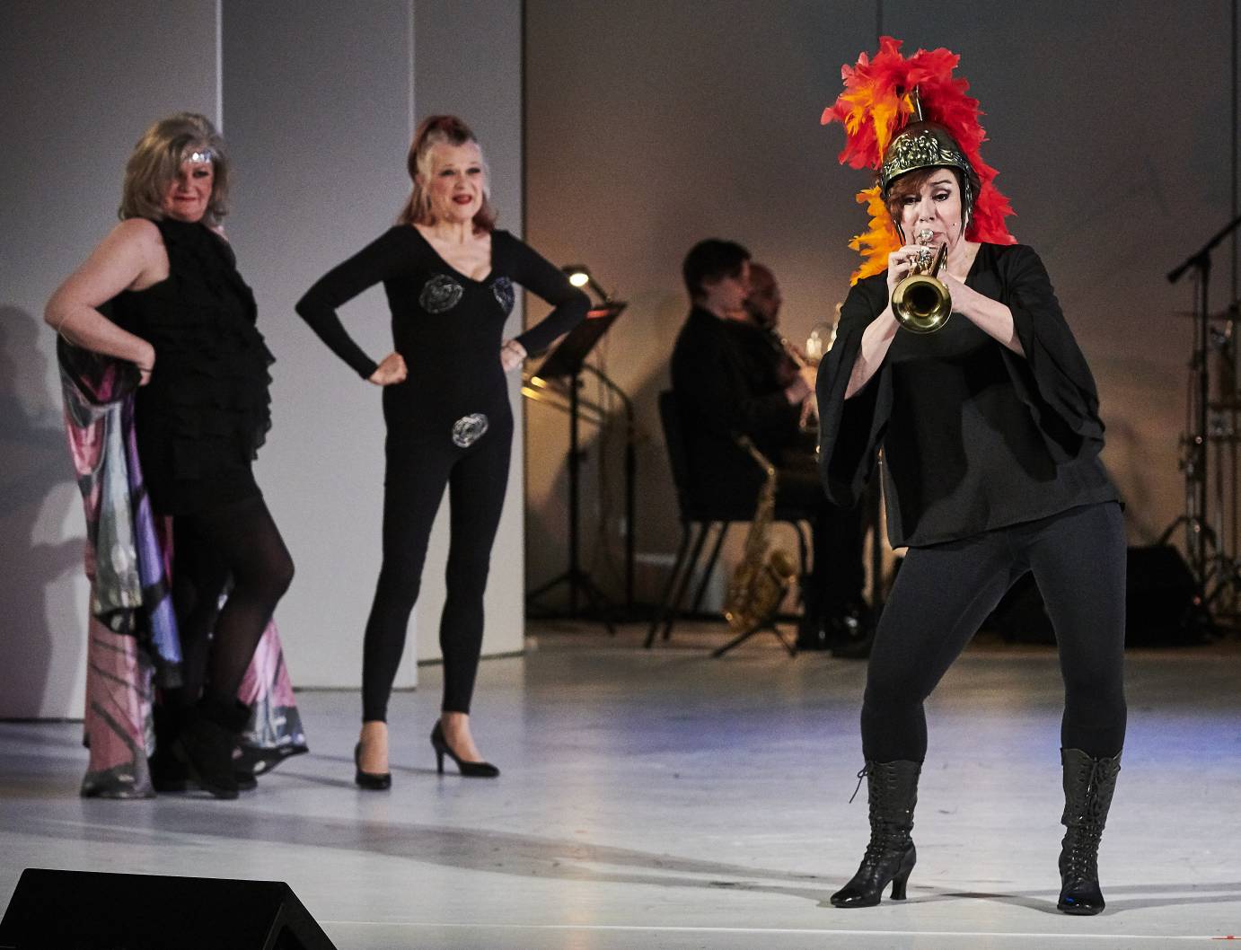 An older lady  blows a trumpet as two sassy middle-aged women watch