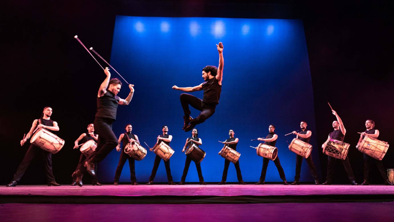 Two men jump, surrounded by a semi circle of men drumming