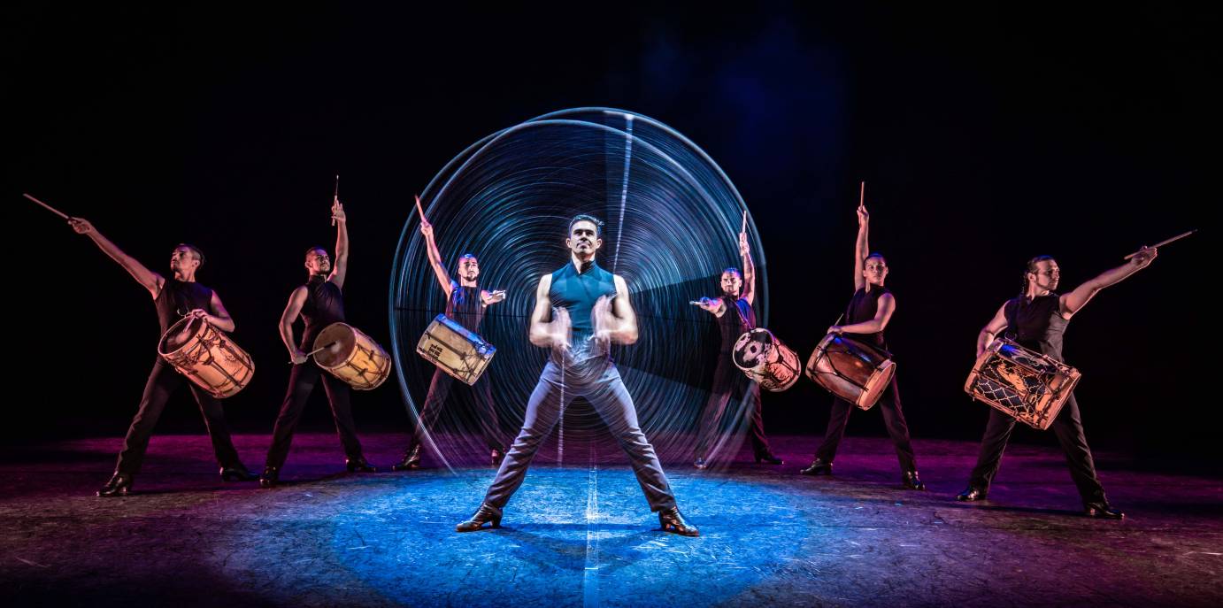 One man twirls a lasso as drummers extend their sticks into the air