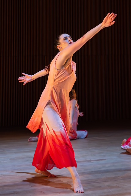 A standing woman in forced-arch, in a long, soft salmon-colored tunic over white and red pants, spirals upward on the diagonal. Her intent face follows her leading arm.