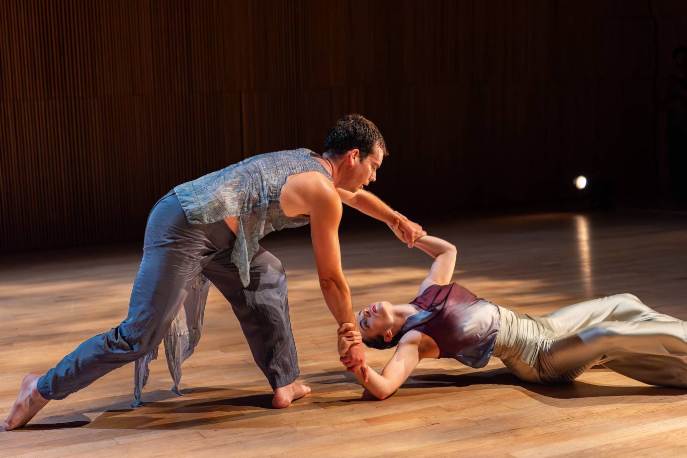 A woman in a shiny beige botton and aubergine top, perched on her right hip, looks into the face of a man, in gray, who holds her forearms.