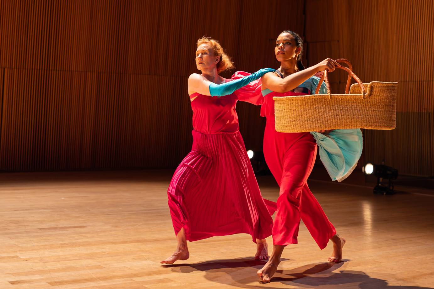 Two women costumed in red and torquoise are caught in mid-jog as they head on the diagonal their faces full of purpose. The downstage woman carries a rush baby bassinet.