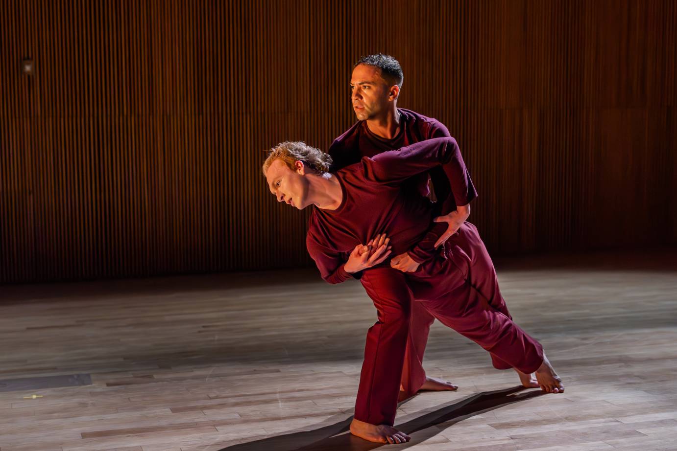 Two men in dark purple long-sleeved tops and pants, on their feet with bodies close, clutch one another. The man in front, in a deep lunge, falls forward, spiraled and looking down, while the man behind holds him up and looks to the distance.