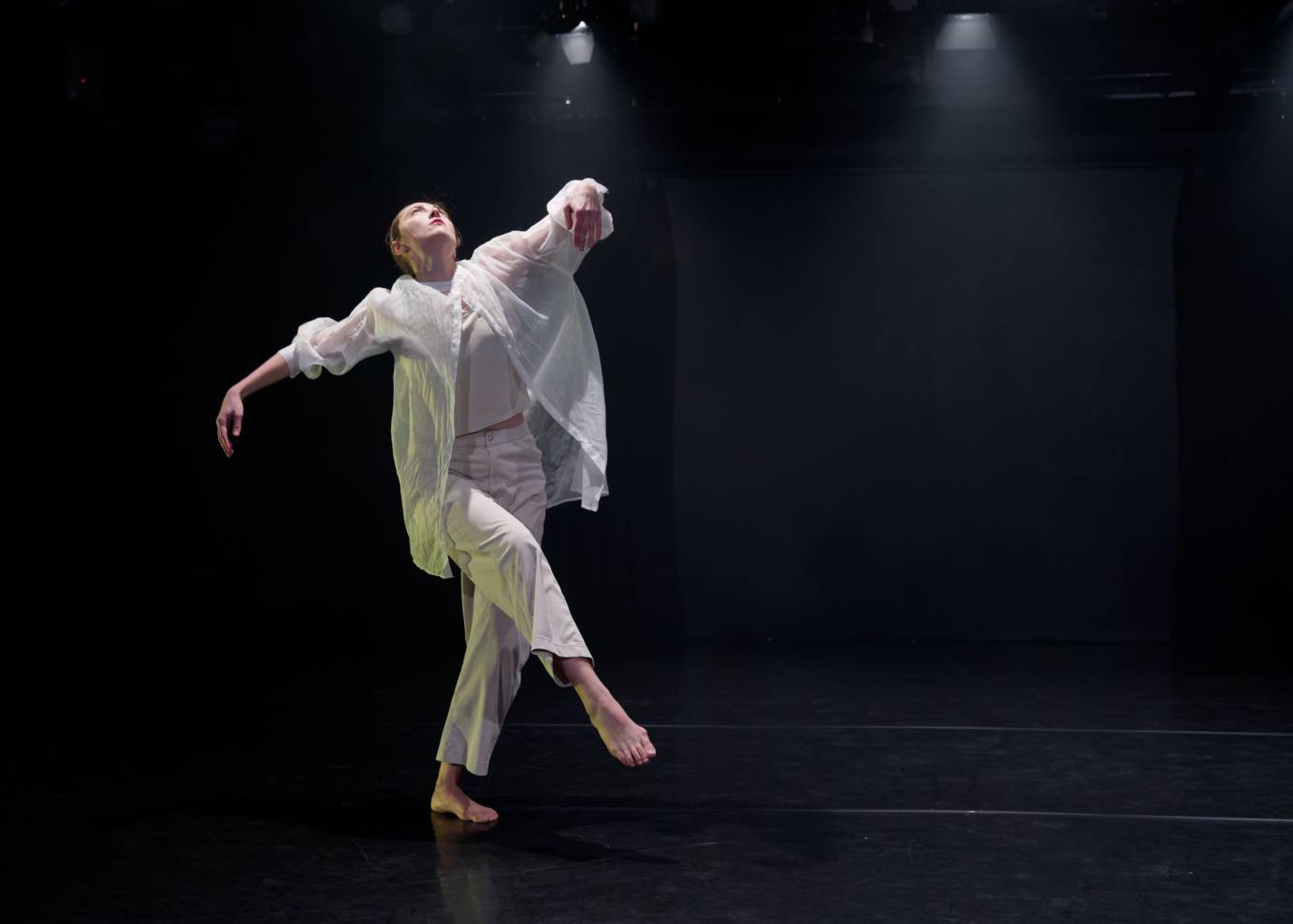 A woman dressed in loose white pants and blouse against a black background softly lifts her right leg and one arm and her face to the ceiling.