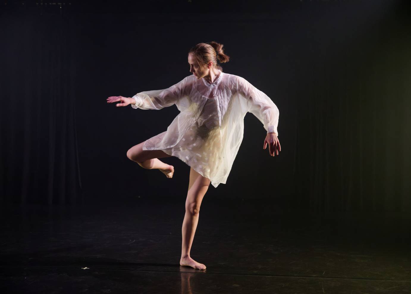 A solo woman with her hair in a French twist gently lifts her right arm balanced on one leg with the other bent behind. She is dressed in a diaphanous knee length tunic.