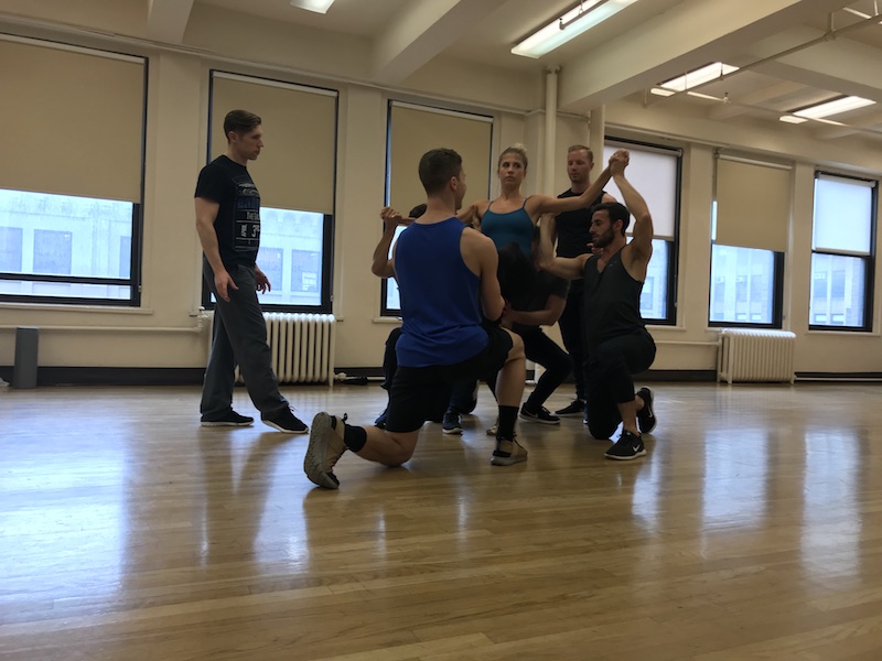 Dancers stand and sit near one another to try to lift a woman overhead.