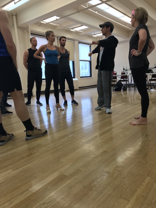 Chris Gattelli in sweats and a baseball cap talks to a group of dancers in a studio.