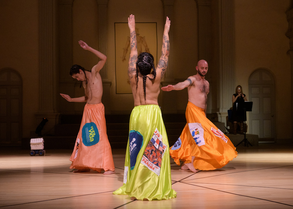 three topless figures dance in a circle. Each artist wears bright neon colored skirts with central American travel slogans patched on to them like Pura Vida or a poster of Panama. Two of the dancers wear long black braids the other, the choreographer is bold with a beard and mustache , the central figure a woman whose back is towards us is covered with beautiful arm tatoos