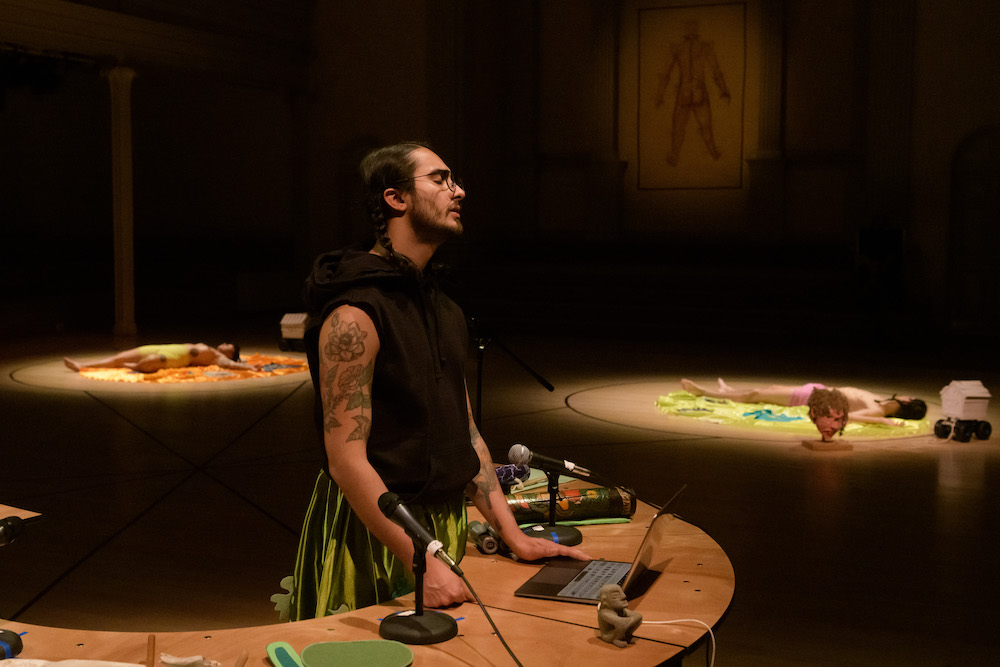 three pools of light on the sanctuary floor at St. Marks. In the closest circle to us a musician stands at a round desk holding mic's and his computer.. a man with long braided black hair, wearing a black sleeveless t and a green skirt, closes his eyes as if in a trance. In two circles of light behind him two figures lie prone