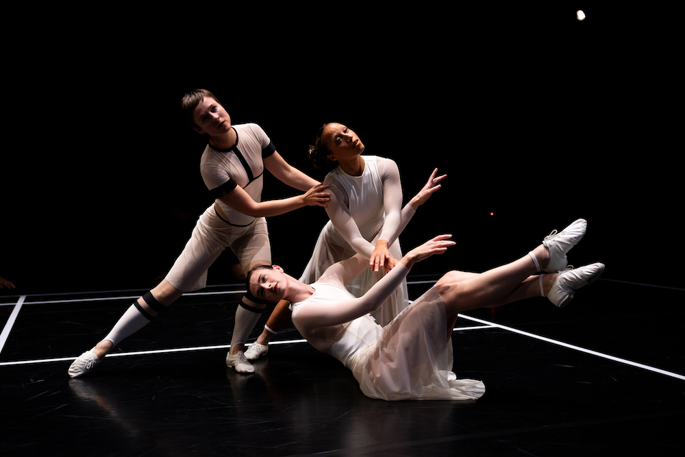 a trio of dancers in variations of white tennis outfits form a triangular tableau...the point is a woman sitting on the ground an above her two other women pose leaning to the right 