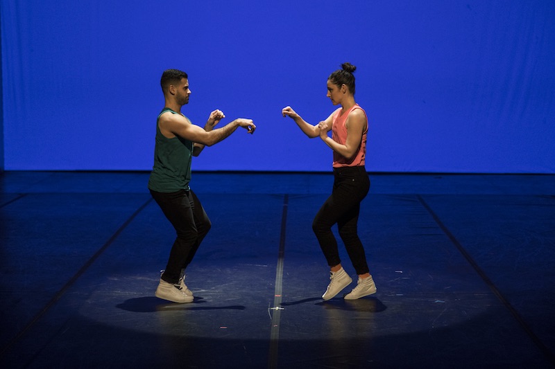Two dancers in bright colored tanks, jeans and white sneakers stand at one another. They both have their hands positioned by paws.