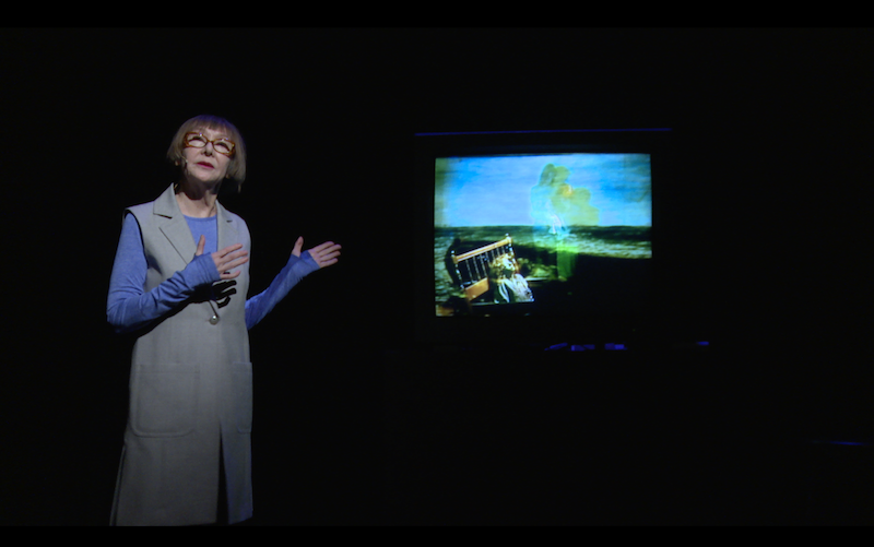 Constance DeJong in a perwinkle top and long grey vest wears classes and points to an old T.V. screen. The screen projects neon blue and yellow colors. Its image is blurry.