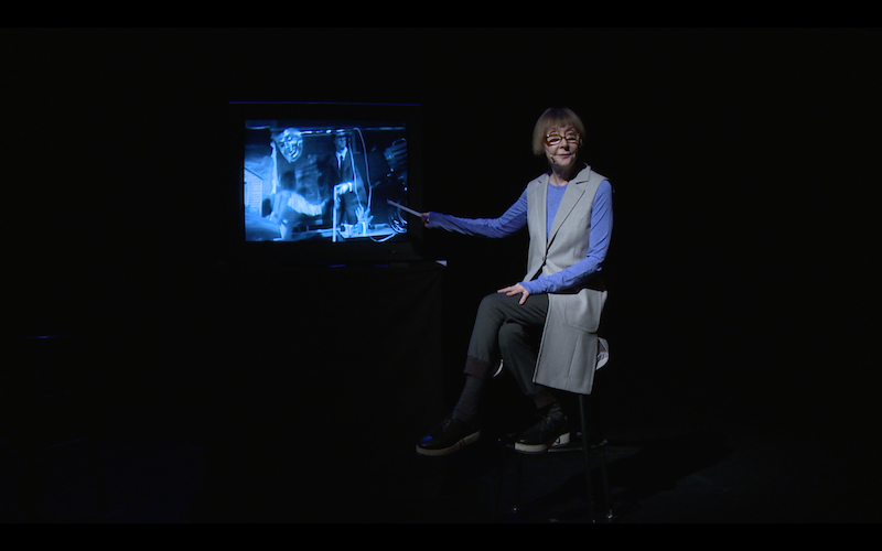 Bespectacled Connie DeJong sits on a stool. Her hand points to a T.V. screen while she looks out to the audience.