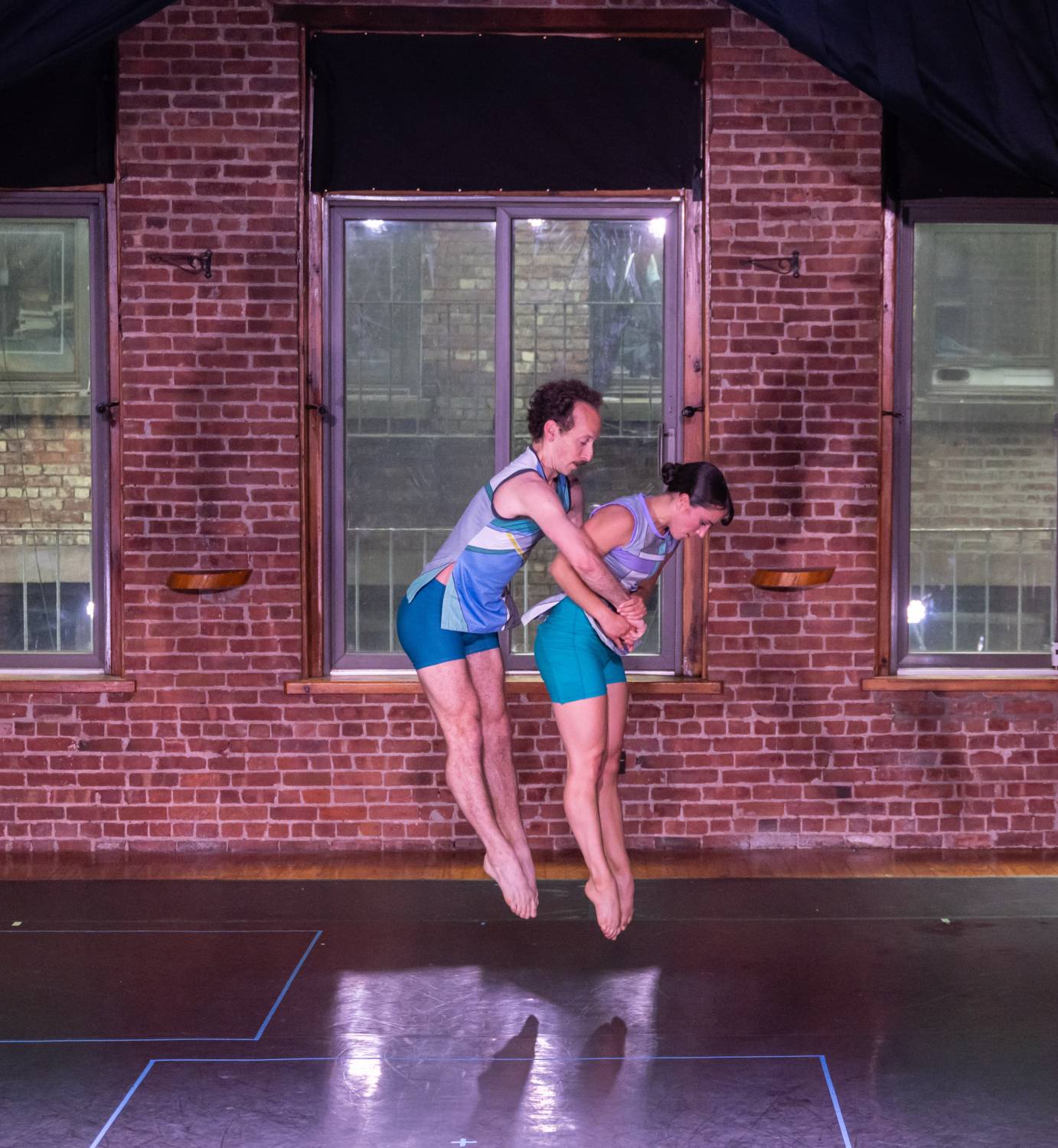 A man behind and a woman jump straight up with legs long. The man clasps his hands around her waist.