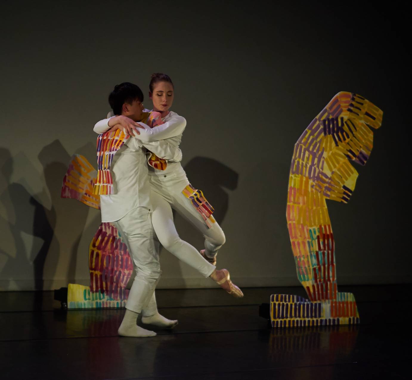 A man with his back to the audience lifts a woman. She is nestled into the side of his body with her arms around his shoulders. They are both costumed in long white pants and shirts with yellow, orange and red woven fabric attached to their costumes at various places. A sculpture in the same colors stands at their height to the right of them.