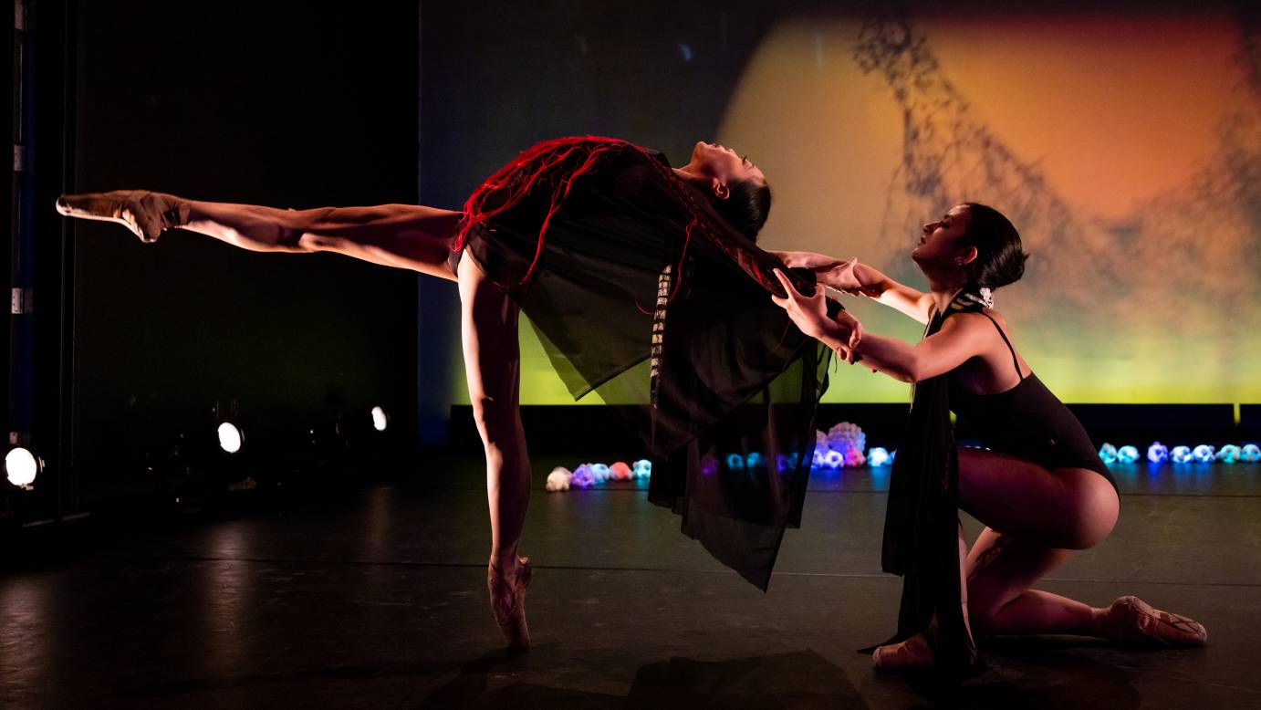 Two barelegged dancers wearing pointe shoes in front of a yellow set. One dancer is kneeling and holding on to the shoulders of the other dancer who is laid out horizontally with leg extended.