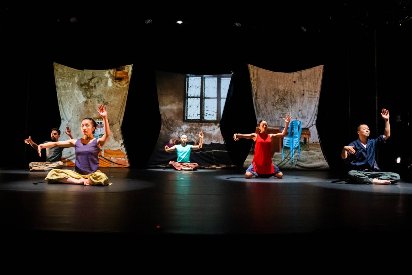 Five dancers sit cross-legged, one arm over their heads, the other extended