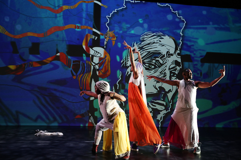 Dancers in gauzy dresses and tunics outstretch their arms. They stand in front of a multicolor background that features the face of a man.