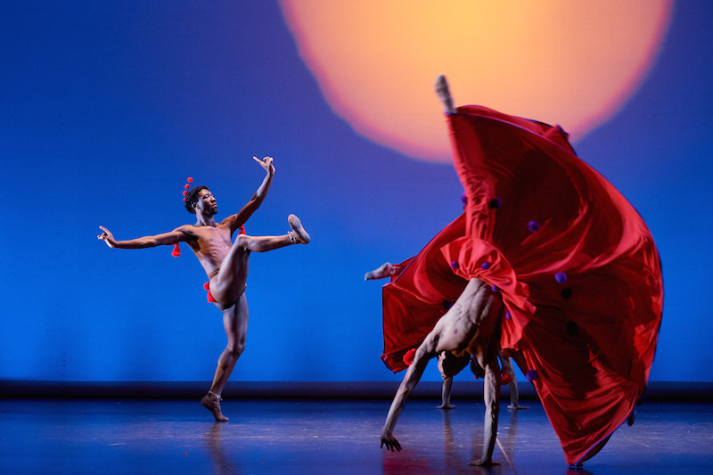 A man flexs his leg in the air in front of him. He's in mid-balance slightly off his center. Another man wears a bright red skirt and is upsidedown in the air.