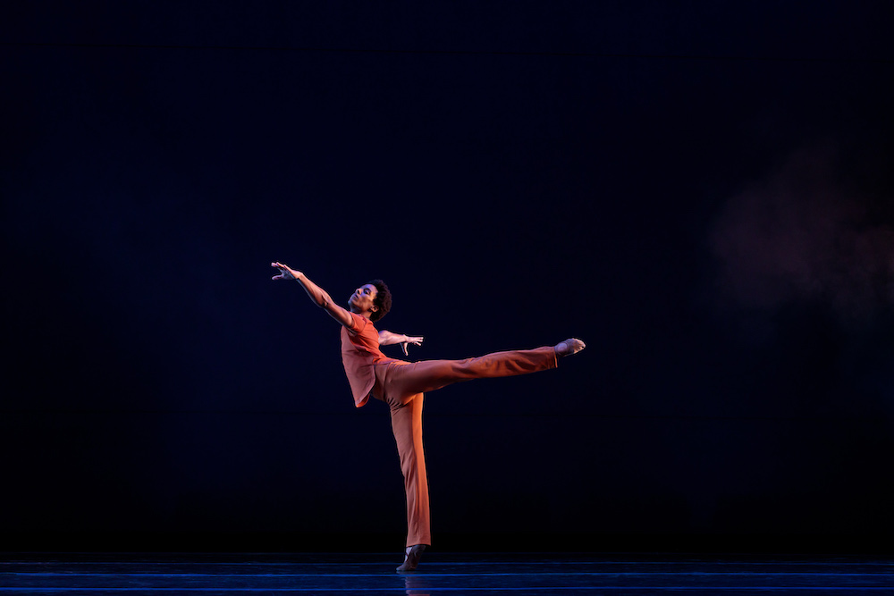 Solo artist Anthony Santos balances in a gorgeous arabesque. He is wearing orange pants and an orange top. 