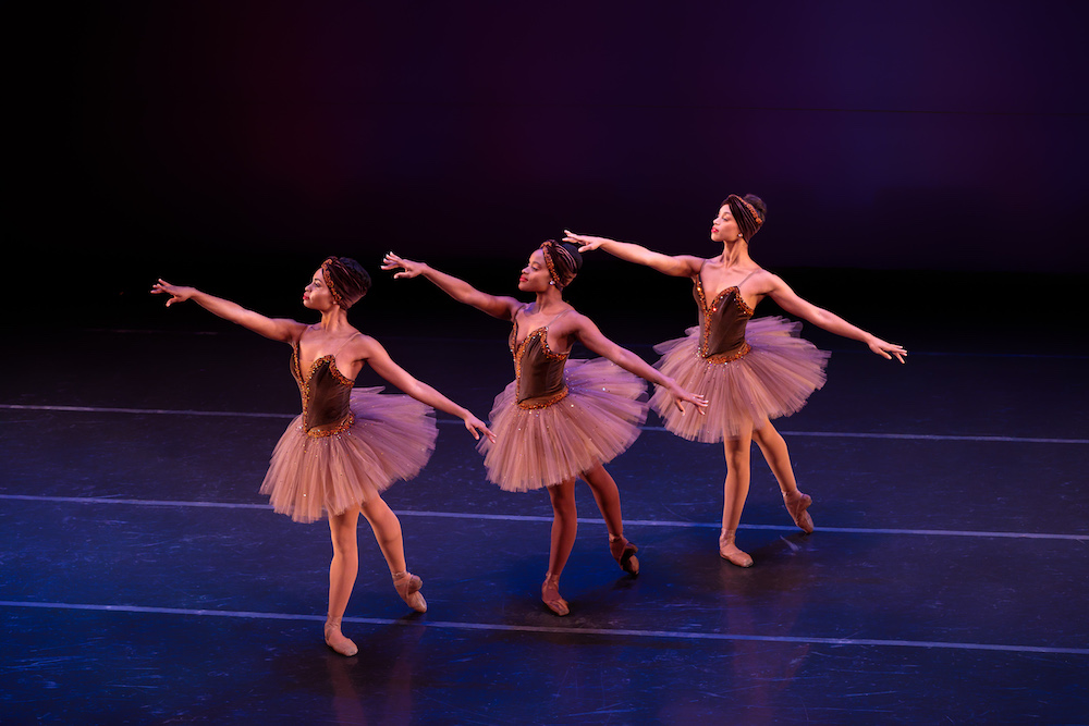 Three Black ballerinas in classical tutus of with crushed velvet and rhinestone bodices and matching crushed velvet head wraps stand in unison, on one leg with the other extended backward on the floor. All different shades, their tights and toe shoes match their various skin tones and they look poised and elegant with their arms out stretched. 