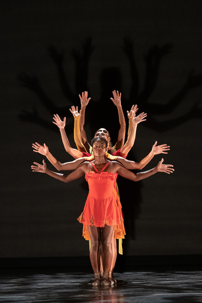 Ingrid Silva wearing bold orange tight bodiced, sleevless dress with a flouncy mini skirt, stands in front of a line of women. We see Silva's entire body but behind her we only notice the other women's arm shapes. 