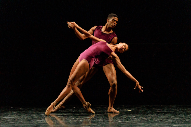 two handsome Black dancers in burgundy leotard costumes partner. the male figure supports his female counterpart as she lunges backwards arching her back and looking out to the audience