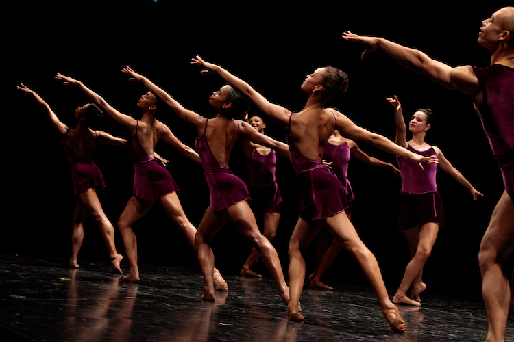 A chorus of male and female dancers pose in an upward facing arabesque postions with both legs on the floor. Donned in  simple burgundy costumes that reflect classroom clothes the women sport nude legs.