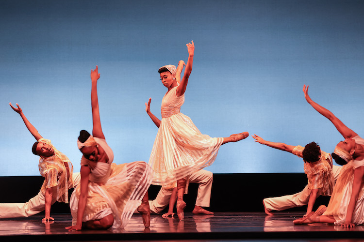 Daphne Lee starring as the Trinidadian born American artist Hazel Scott lifts her arms high as she balances on one leg with the other bent behind her.  the company kneels on the floor surrounding her. All wear ivory colored theatrical approximations of West Indian dress