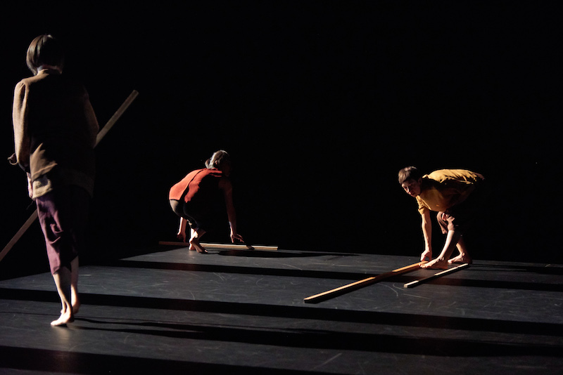 Three women in semi darkness hold sticks or lay down the long wooden rods. They cast long shadows.