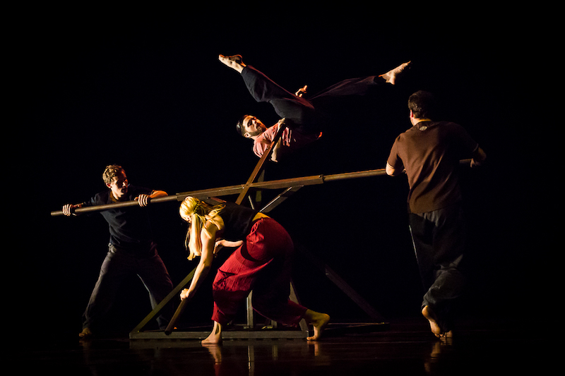 Four dancers surround a geometrically structured wood object. One dancer is in midst of hoisting himself up onto one of the structure's spindles.