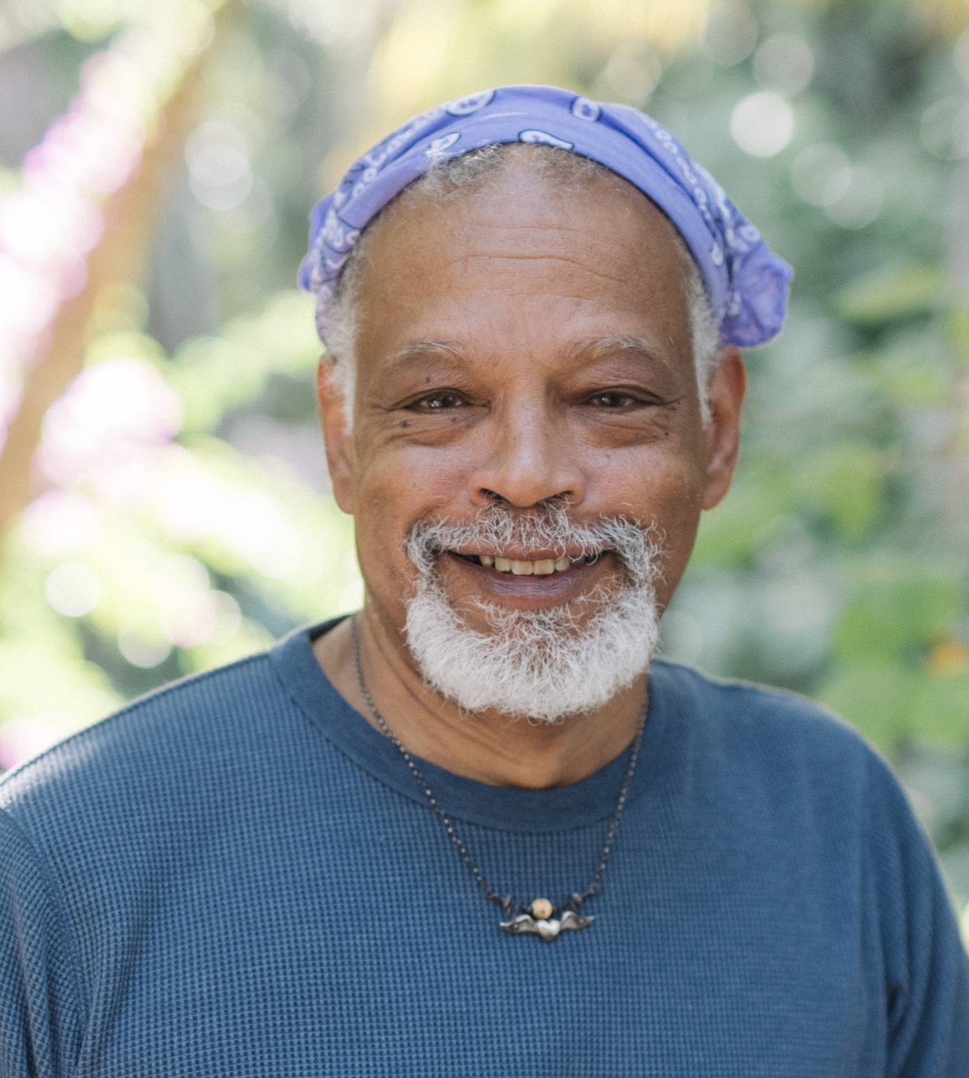 portrait of a black male artist Ishmael Houston- Jones, with tufts of black grey hair poking out from a purple and white head scarf, white beard , big smile, blue flannel shirt and a beaded necklace 
