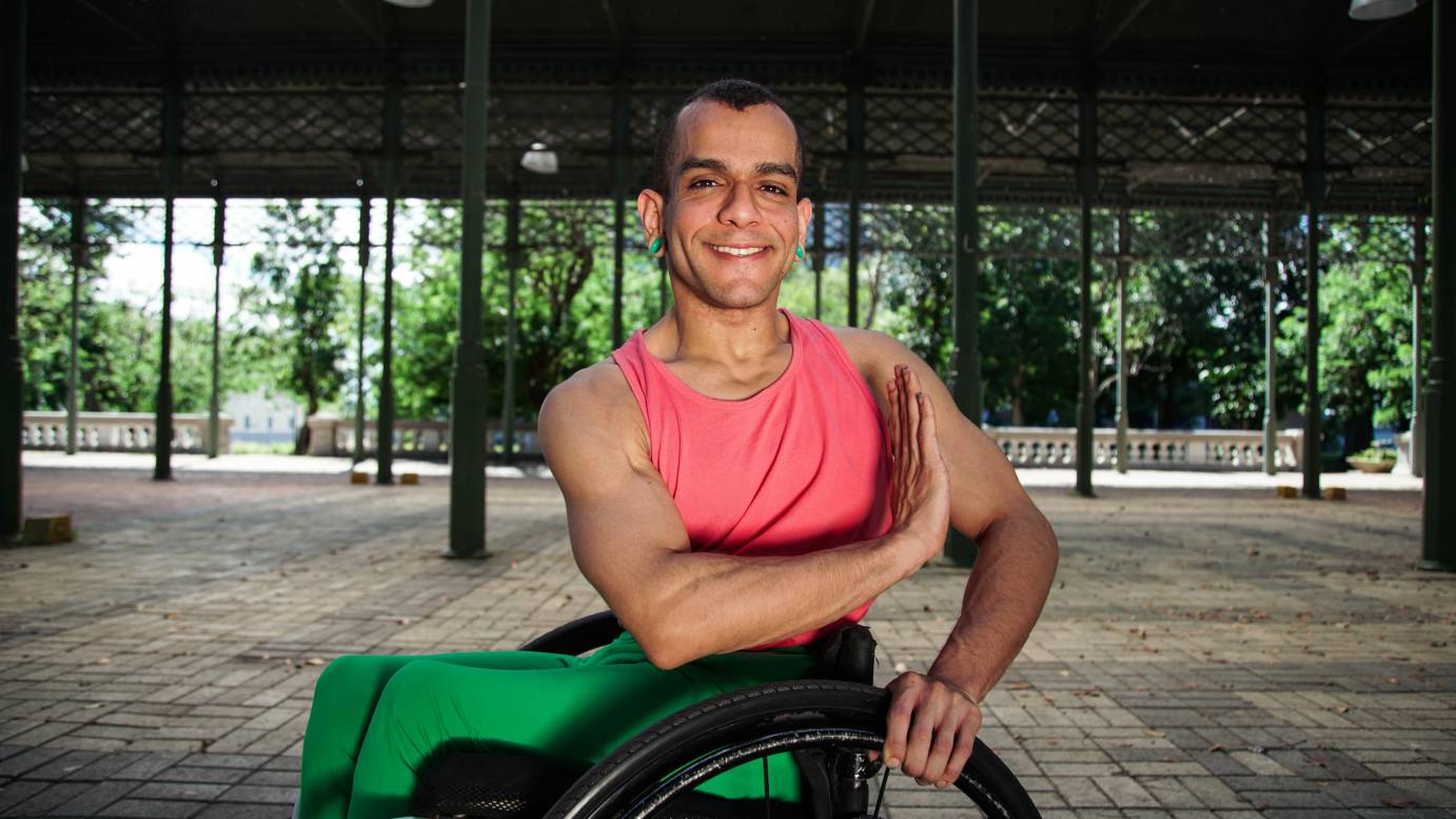 JanpiStar portrait of artist under a bridge, brown skin, smiling, pink red sleeveless shirt, and green pans , sitting on a wheel chair, nature in background, green earrings