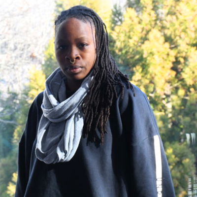 Queer Black artist Ni'Ja Whitson, with long braids a dark blue shirt and a light blue scarf, a nose ring set against a green tree back ground