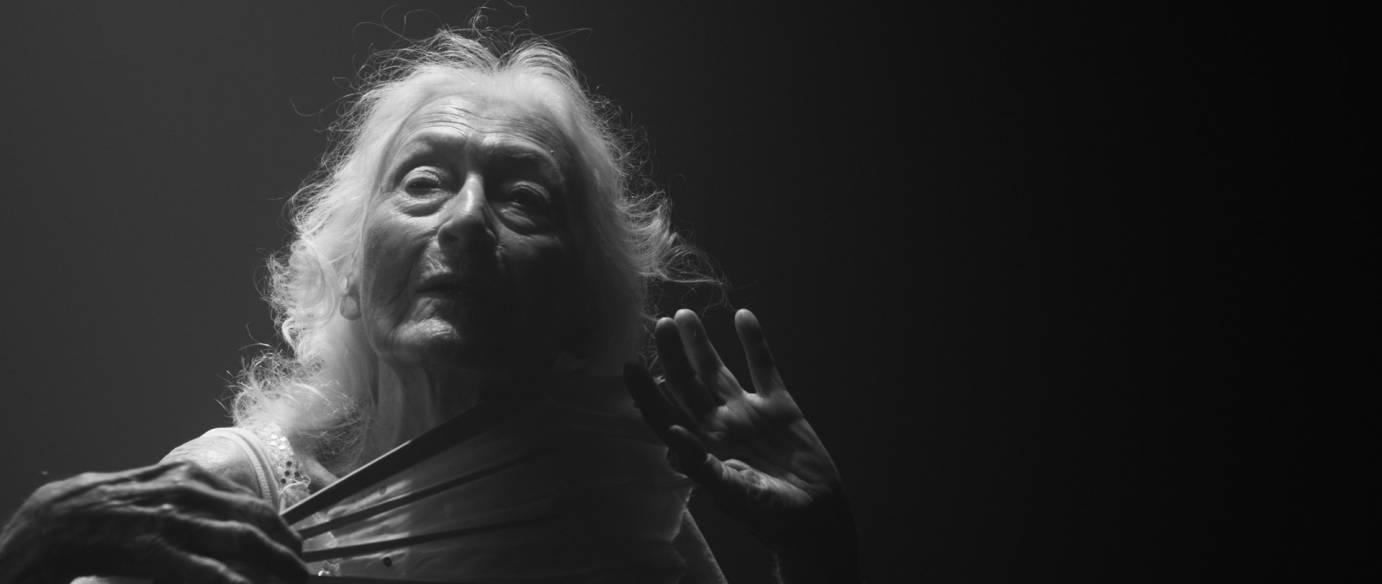 a 100 plus year old dancer with flowing white hair looks out into a dream like world, holding a fan in one hand with her other hand expressively drawn back, the lighting accentuates the beauty of her white hair and wrinkled face