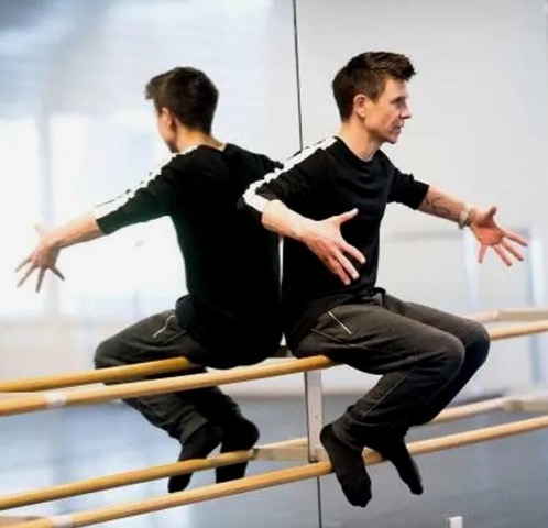 white man in black outfit with white stripe in a dance studio sitting on the barre