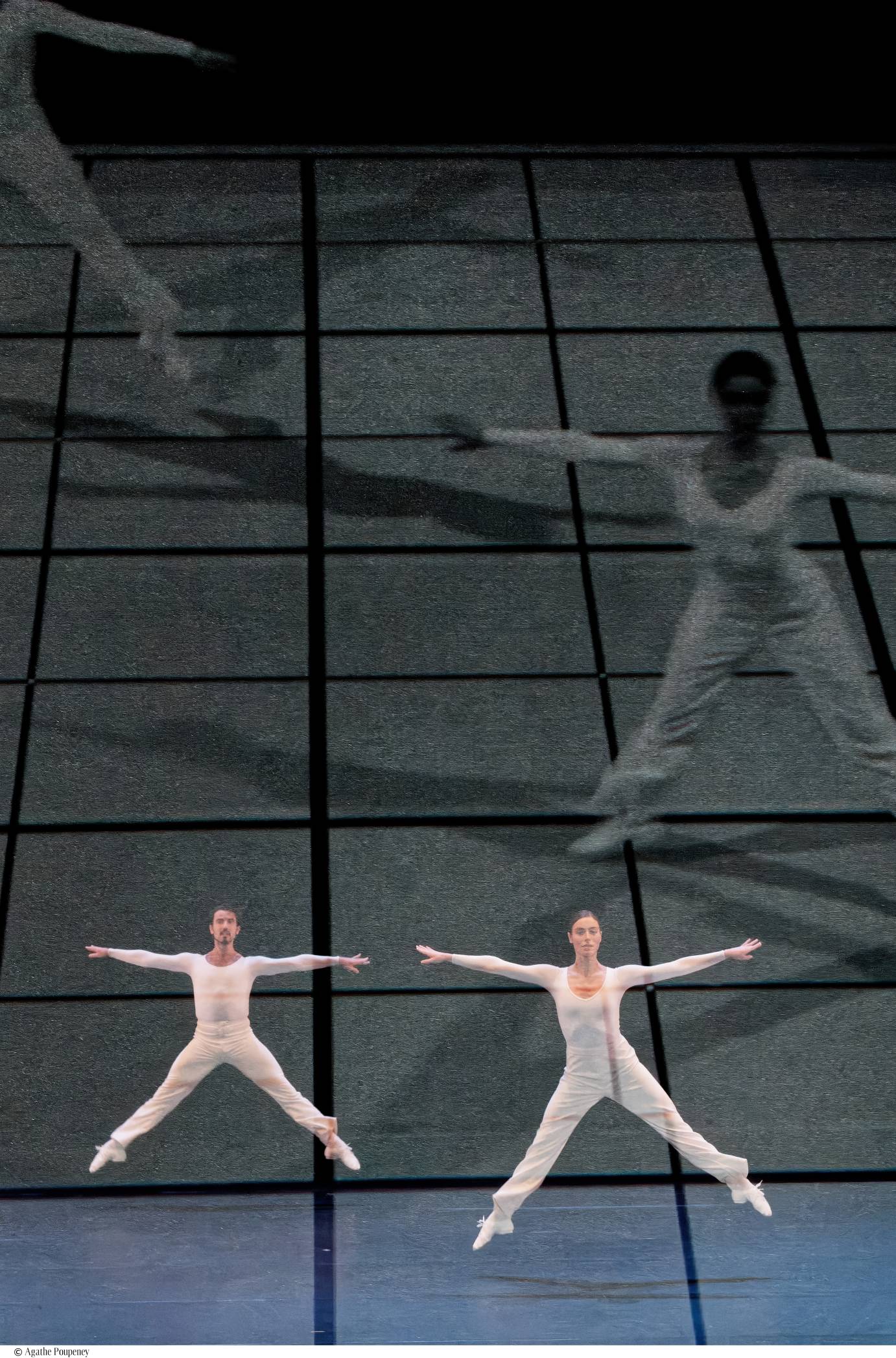 Pair of white, long-sleeved unitard dancers facing downstage, jumping in a wide second position, arms horizontal. Their larger-than-life images are projected simultaneously.