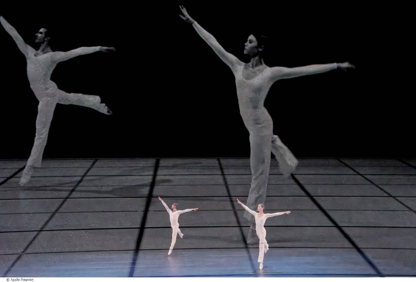 Live duet, woman and man dressed in white long-sleeved unitards overlayed by film, dancers dancing same steps on a white with black grid, dressed in same unitards, behind live dancers.