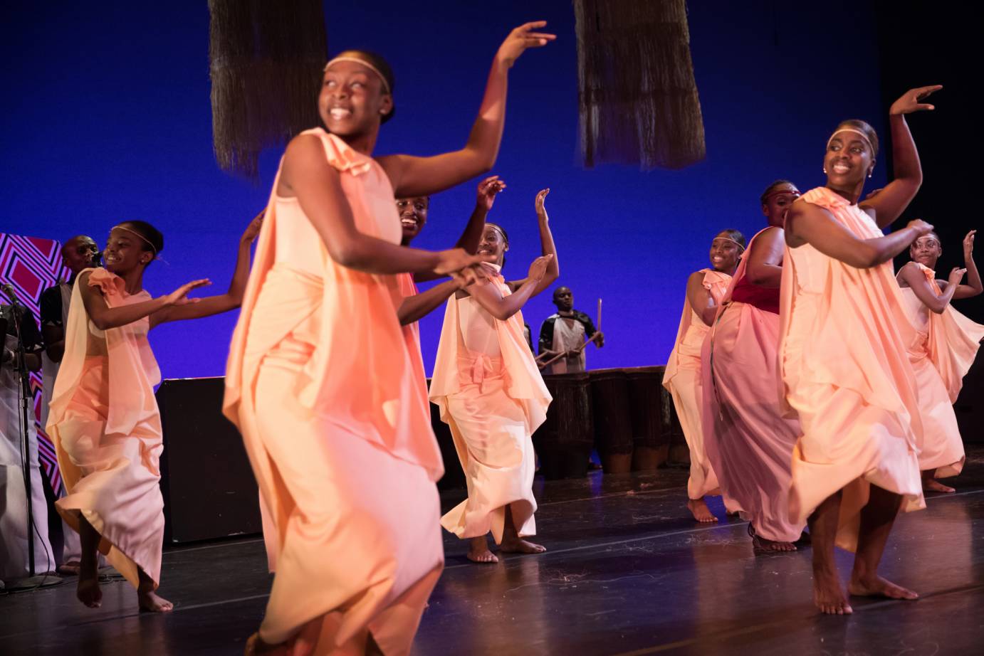 A group of peach-clad women smile in profile as they lift one leg in a step, arms extended 